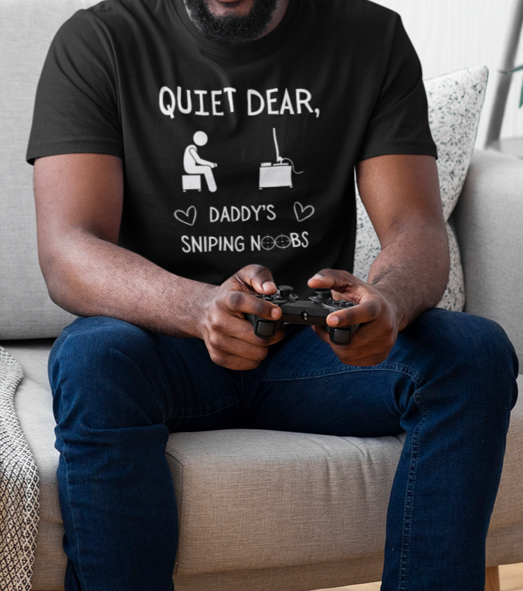 A model wearing a black t-shirt that reads Quiet Dear, Daddy's Sniping Noobs in white text. The lower text is framed by two hearts, and the O's are in the shape of sniper scopes. In the center of the shirt is an image of a person holding a controller sitting across from a TV.