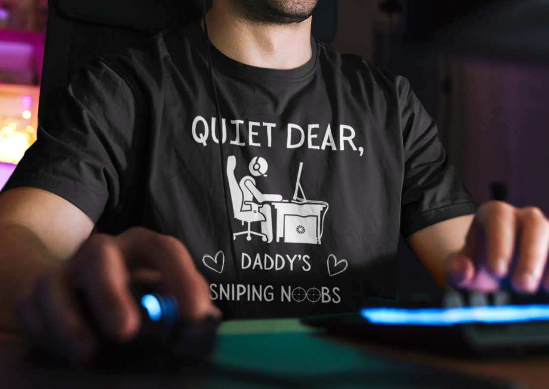 A model wearing a black t-shirt that reads Quiet Dear, Daddy's Sniping Noobs in white text. The lower text is framed by two hearts, and the O's are in the shape of sniper scopes. In the center of the shirt is an image of a person at a desk with a gaming PC.