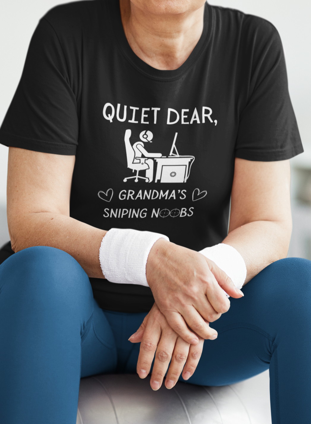 A model wearing a black t-shirt that reads Quiet Dear, Grandma's Sniping Noobs in white text. The lower text is framed by two hearts, and the O's are in the shape of sniper scopes. In the center of the shirt is an image of a person at a desk with a gaming PC.